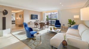Una moderna sala de estar inspirada en el Hotel The One Barcelona GL, con un sofá blanco, sillas azules y una mesa de comedor junto al gran ventanal. Plantas exuberantes y obras de arte cautivadoras completan la decoración, reflejando el elegante estilo del hotel. - ForTwo - Planes para parejas