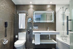 El baño del Hotel Vincci Gala de Barcelona cuenta con un lavabo de pared, un gran espejo, una bañera, una ducha y azulejos con textura oscura. - ForTwo - Planes para parejas