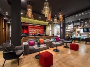 El Hotel Vincci Gala de Barcelona cuenta con un moderno salón con cómodos asientos, cojines coloridos e iluminación contemporánea. Un elegante bar complementa las vibrantes estanterías rojas, representando el ambiente chic de la ciudad. - ForTwo - Planes para parejas