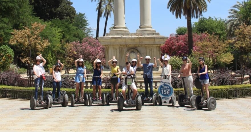 Un grupo posa en Segways durante la pintoresca "Ruta en Segway por Sevilla" en un parque, con palmeras y columnas al fondo. - ForTwo - Planes para parejas