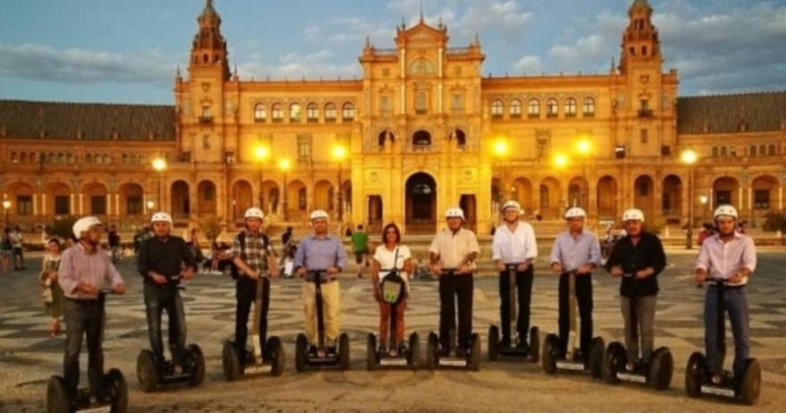 Un grupo disfruta del aura encantadora de Sevilla iluminada al anochecer en la 'Ruta en Segway por Sevilla', deslizándose junto a un edificio histórico bellamente iluminado. - ForTwo - Planes para parejas