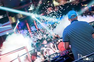 En SHÔKO Barcelona, un DJ con gorra azul y camisa a rayas anima la atmósfera de la discoteca mientras llueve confeti sobre la vibrante multitud. - ForTwo - Planes para parejas