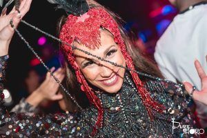 En una fiesta animada, alguien irradia alegría con un traje de lentejuelas y un tocado de plumas rojas que sostiene cordones de cuentas, evocando las glamurosas noches de SHÔKO Barcelona. - ForTwo - Planes para parejas