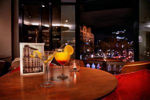 Dos cócteles se sientan en una mesa redonda en el Wilson Boutique Hotel (Barcelona), con las luces centelleantes de la ciudad filtrándose a través de grandes ventanales y proyectando un cálido resplandor en la habitación tenuemente iluminada. - ForTwo - Planes para parejas
