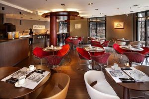 En el Wilson Boutique Hotel (Barcelona), un moderno restaurante con sillas rojas y blancas y mesas preparadas ofrece impresionantes vistas a través de grandes ventanales que permiten a los comensales observar a la gente explorar el buffet adyacente. - ForTwo - Planes para parejas