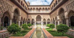 Descubra el encantador patio del Real Alcázar de Sevilla, con sus intrincados arcos, exuberante vegetación y una estrecha alberca central. Planifique su "Visita al Real Alcázar de Sevilla sin esperas" para sumergirse por completo en esta maravilla arquitectónica bajo un cielo parcialmente nublado. - ForTwo - Planes para parejas