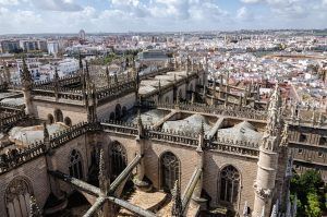 El producto "Visita la Catedral de Sevilla sin esperas" ofrece impresionantes vistas aéreas de la majestuosa Catedral de Sevilla, que se alza orgullosa en medio del bullicioso paisaje urbano bajo un espectacular cielo nublado. - ForTwo - Planes para parejas