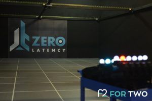 En una habitación con poca luz y suelo cuadriculado, el logotipo de la Experiencia de realidad virtual multijugador Zerolatency (Terrassa) destaca en la pared, prometiendo emocionantes aventuras. El equipo borroso en primer plano invita a los jugadores a sumergirse en otra dimensión. - ForTwo - Planes para parejas