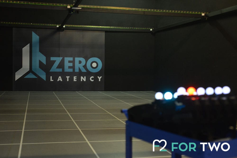 En una habitación con poca luz y suelo cuadriculado, el logotipo de la Experiencia de realidad virtual multijugador Zerolatency (Terrassa) destaca en la pared, prometiendo emocionantes aventuras. El equipo borroso en primer plano invita a los jugadores a sumergirse en otra dimensión. - ForTwo - Planes para parejas