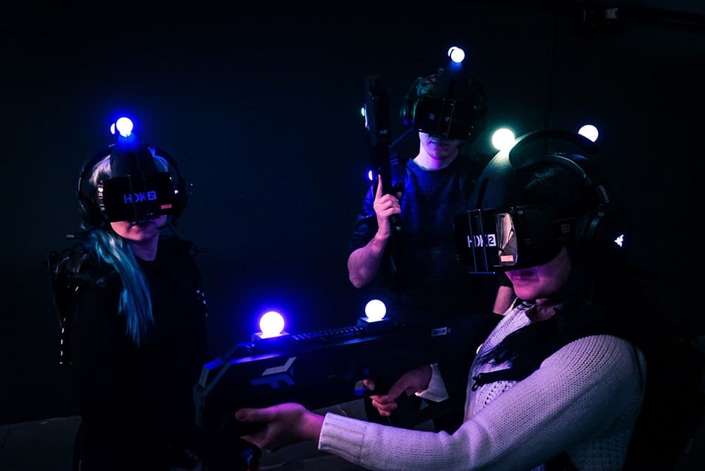 Tres personas con gafas de realidad virtual y controladores con marcadores iluminados se sumergen en emocionantes aventuras en una habitación oscura con la Experiencia de realidad virtual multijugador Zerolatency (Terrassa). - ForTwo - Planes para parejas