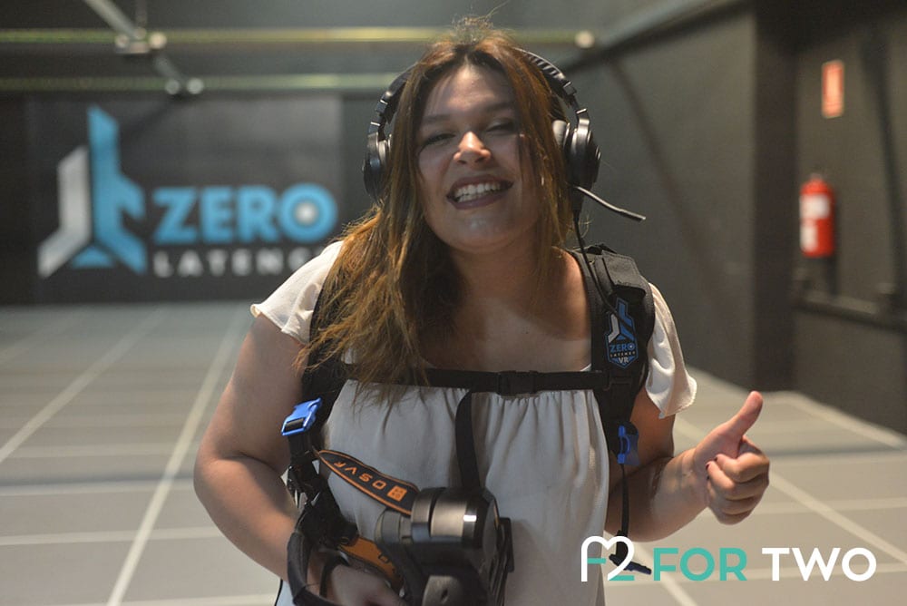 En la arena Zerolatency VR, una persona sonríe con el pulgar hacia arriba, equipada con equipo y casco, mientras el logotipo de Zero Latency brilla detrás de ella, encapsulando la esencia emocionante de la Experiencia de realidad virtual multijugador Zerolatency en Terrassa. - ForTwo - Planes para parejas