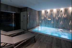 Relájese con estilo en el Hotel Catalonia Magdalenes de Barcelona con un moderno jacuzzi interior con agua iluminada en azul, tumbonas y paneles de pared con textura oscura. Disfrute de la comodidad y haga de su estancia algo inolvidable. - ForTwo - Planes para parejas