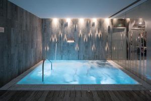 En el Hotel Catalonia Magdalenes (Barcelona), la piscina cubierta ofrece una iluminación tenue y una barandilla metálica. Los chorros de agua crean relajantes ondas, mientras que las paredes revestidas de madera y una puerta de cristal realzan su ambiente sereno, convirtiéndola en un oasis urbano. - ForTwo - Planes para parejas