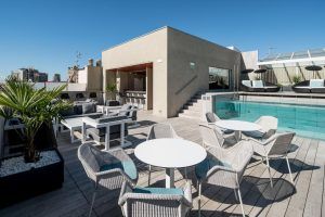 El patio de la azotea del Hotel Catalonia Magdalenes (Barcelona) exhibe muebles de mimbre, mesas redondas, una piscina, tumbonas y un encantador edificio contra el cielo azul claro. - ForTwo - Planes para parejas