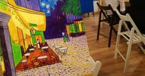 Una pintura colorida sobre un caballete captura una escena de café con sillas plegables, evocando la esencia vibrante de Madrid, que recuerda a un Taller creativo de pintura de cuadros. - ForTwo - Planes para parejas