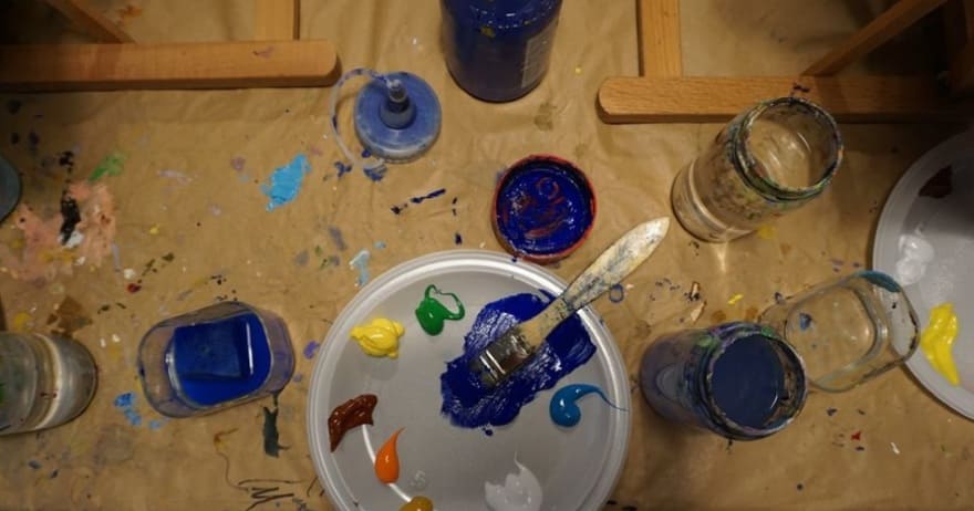 Varios botes de pintura y una paleta con un pincel reposan sobre una superficie desordenada y salpicada de pintura, similar al vibrante Taller creativo de pintura de cuadros de Madrid. - ForTwo - Planes para parejas