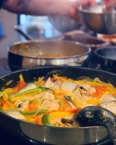 Primer plano de pescado y verduras chisporroteando en una sartén bajo un cucharón, evocando una animada escena del Taller de cocina en Sants con Taller 24 (Barcelona). Al fondo, desenfocado, otra olla burbujea junto a la figura del capataz que atiende esta sabrosa creación. - ForTwo - Planes para parejas