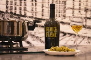 Una botella de vino y una copa se ubican elegantemente junto a una olla y aceitunas en una mesa, evocando la escena culinaria de Taller de cocina en Sants con Taller 24 en Barcelona, todo sobre un llamativo fondo blanco y negro. - ForTwo - Planes para parejas