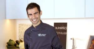 Un chef sonriente se encuentra en la cocina de Taller de Cocina en Illa Diagonal (Barcelona), vistiendo un uniforme oscuro y con productos frescos dispuestos de forma atractiva en el mostrador detrás de él. - ForTwo - Planes para parejas