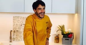 Un chef con abrigo amarillo sonríe en una bulliciosa cocina en Illa Diagonal, rodeado de verduras frescas y herramientas, listo para dirigir el atractivo "Taller de cocina en Illa Diagonal (Barcelona). - ForTwo - Planes para parejas