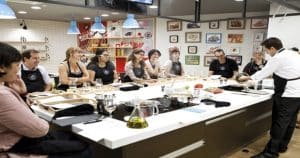 En el Taller de cocina en Illa Diagonal de Barcelona, un chef demuestra con maestría técnicas de cocina a estudiantes atentos reunidos alrededor de una isla de cocina. - ForTwo - Planes para parejas