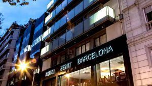 Vista de la calle de la moderna fachada del Hotel Zenit Barcelona con un cartel iluminado al anochecer, donde los huéspedes pueden disfrutar de una espaciosa habitación doble, disfrutar de un desayuno buffet y ver un espectáculo de flamenco antes de explorar los edificios cercanos. - ForTwo - Planes para parejas