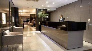 Recepción del hotel elegantemente iluminada con una persona en el escritorio y una zona de estar a la izquierda, perfecta para comenzar tu Escapada con habitación doble, desayuno buffet y espectáculo de flamenco (Barcelona). - ForTwo - Planes para parejas