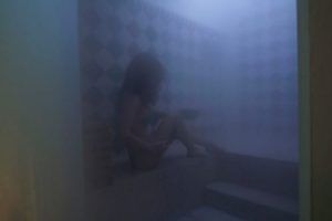 En medio de la niebla que recuerda a un hammam tradicional, alguien se sienta en un escalón de baldosas, parcialmente oculto por la niebla, evocando la serenidad atemporal del Hammam con masaje de 15 minutos en los baños árabes de Al-Haram en Granada. - ForTwo - Planes para parejas