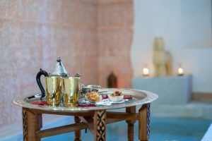 Un juego de té tradicional con aperitivos espera en una mesa de madera junto a una piscina reluciente, rodeada de velas y una estatua de piedra, evocando la tranquilidad del "Hammam privado en pareja en los baños árabes de Al-Haram (Granada). - ForTwo - Planes para parejas