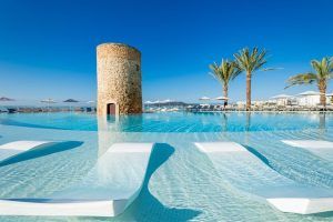 Disfrute de la impresionante piscina infinita del Hotel Torre del Mar con tumbonas sumergidas, ubicada junto a una encantadora torre de piedra y rodeada de elegantes palmeras bajo el cielo azul claro de Ibiza. - ForTwo - Planes para parejas