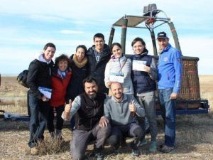 Un grupo sonríe de alegría, posando junto a una cesta de globo aerostático en un campo frondoso, listos para su emocionante aventura con la "Oferta: vuelo en globo con hotel en Segovia". - ForTwo - Planes para parejas