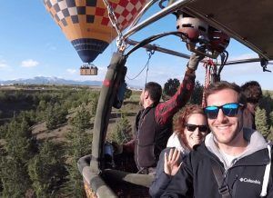 Un grupo sonriente en un globo aerostático sobrevuela los pintorescos paisajes de Segovia con otro globo y montañas detrás de ellos, disfrutando de una experiencia inolvidable con la "Oferta: vuelo en globo con hotel en Segovia" antes de relajarse en su hotel. - ForTwo - Planes para parejas