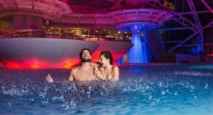 Una pareja disfruta de un baño nocturno en el Spa Caldea de Andorra, rodeados de luces de colores y una fuente de agua futurista, con el paquete "Caldea Spa + Comida o cena para dos". - ForTwo - Planes para parejas