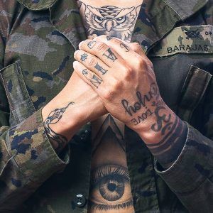 Una persona con una chaqueta de camuflaje adornada con un parche de BARAJAS y tatuajes de un búho en los dedos y el pecho irradia la esencia vanguardista capturada durante la sesión de fotografía de pareja de estudio en Madrid. Las manos entrelazadas reflejan la atmósfera única del lugar. - ForTwo - Planes para parejas