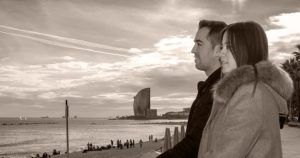 Una pareja contempla el mar desde un paseo marítimo de Barcelona, con el paisaje urbano y las nubes detrás, capturando la esencia de la Sesión de fotografía romántica al aire libre (Barcelona). - ForTwo - Planes para parejas