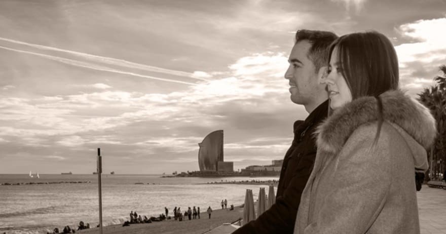 Una pareja contempla el mar desde un paseo marítimo de Barcelona, con el paisaje urbano y las nubes detrás, capturando la esencia de la Sesión de fotografía romántica al aire libre (Barcelona). - ForTwo - Planes para parejas