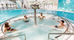 Cuatro personas se relajan en un jacuzzi circular con chorros, situado en la luminosa zona de piscina cubierta del prestigioso "Caldea Spa + Comida o cena para dos" de Andorra. - ForTwo - Planes para parejas
