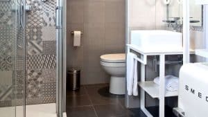 Un moderno baño, diseñado al estilo elegante del Hotel Ryans La Marina, cuenta con azulejos grises, ducha, lavabo, inodoro y toallero. Este espacio es la manera ideal de empezar el día con Escapada Hotel Ryans La Marina con desayuno incluido en Ibiza. - ForTwo - Planes para parejas