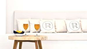 Dos vasos de cerveza y un bol de patatas fritas descansan sobre una mesa de madera frente a un sofá blanco con almohadas R, capturando la atmósfera relajada del Hotel Escapada Ryans La Marina con desayuno incluido (Ibiza). - ForTwo - Planes para parejas