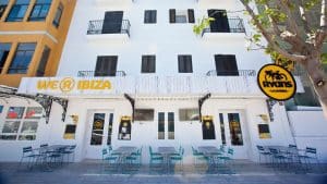La cafetería al aire libre cuenta con sillas y mesas azules, frente a un edificio blanco impecable con el cartel de WE [R] IBIZA y el logotipo de Ryans Lab. Con una ubicación ideal cerca del Escapada Hotel Ryans La Marina con desayuno incluido (Ibiza), es ideal para un desayuno relajado. - ForTwo - Planes para parejas