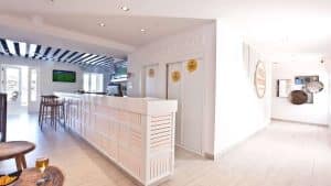 La recepción del Escapada Hotel Ryans La Marina con desayuno incluido (Ibiza) cuenta con un elegante escritorio blanco, taburetes, televisor y arte decorativo en la pared. Su diseño luminoso y limpio refleja el ambiente animado de Ibiza. Disfruta del desayuno incluido para empezar el día de la mejor manera. - ForTwo - Planes para parejas