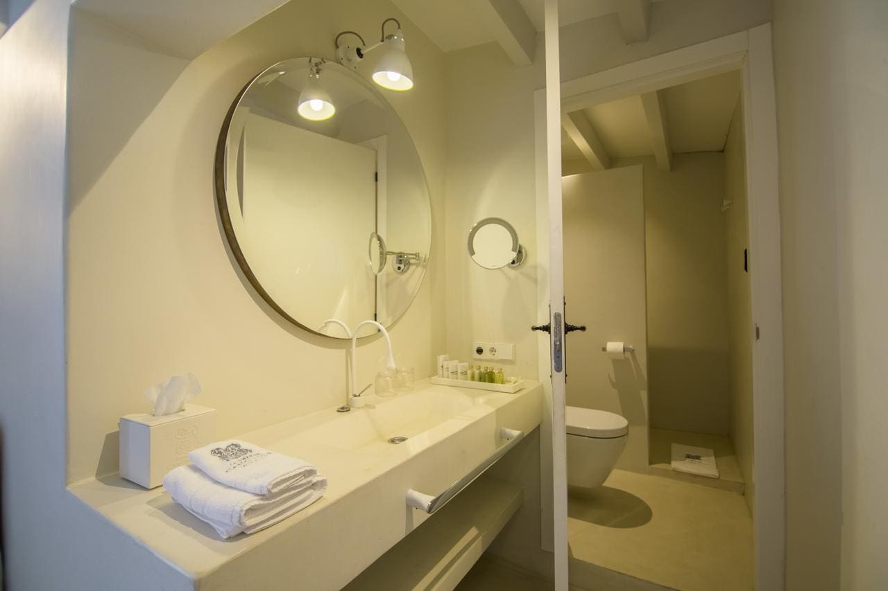 El baño minimalista del Hotel Torre del Canónigo, Ibiza incluye un espejo redondo, un lavabo elegante, lujosas toallas, artículos de tocador esenciales y un inodoro en tonos neutros relajantes. - ForTwo - Planes para parejas