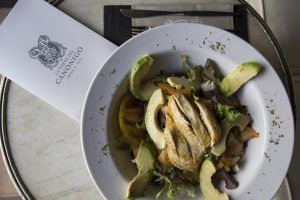 Una vibrante ensalada con rodajas de aguacate, pollo y verduras se presenta elegantemente en un plato blanco junto a un menú y cubiertos en el Hotel Torre del Canónigo (Ibiza). - ForTwo - Planes para parejas