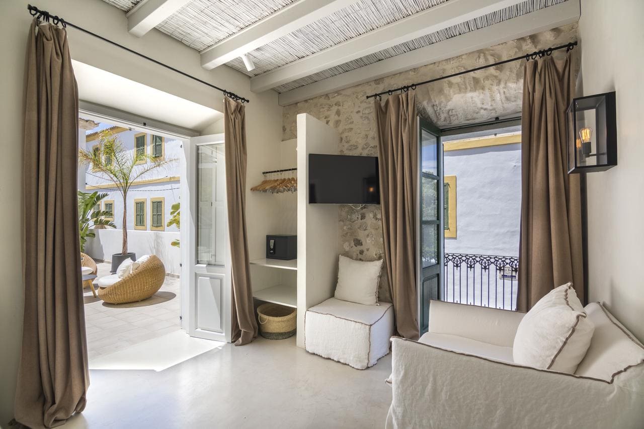 Una acogedora habitación del Hotel Torre del Canónigo (Ibiza) cuenta con paredes blancas, un sofá, puertas de acceso al patio y un televisor montado en la pared; la luz natural llena el espacio. - ForTwo - Planes para parejas