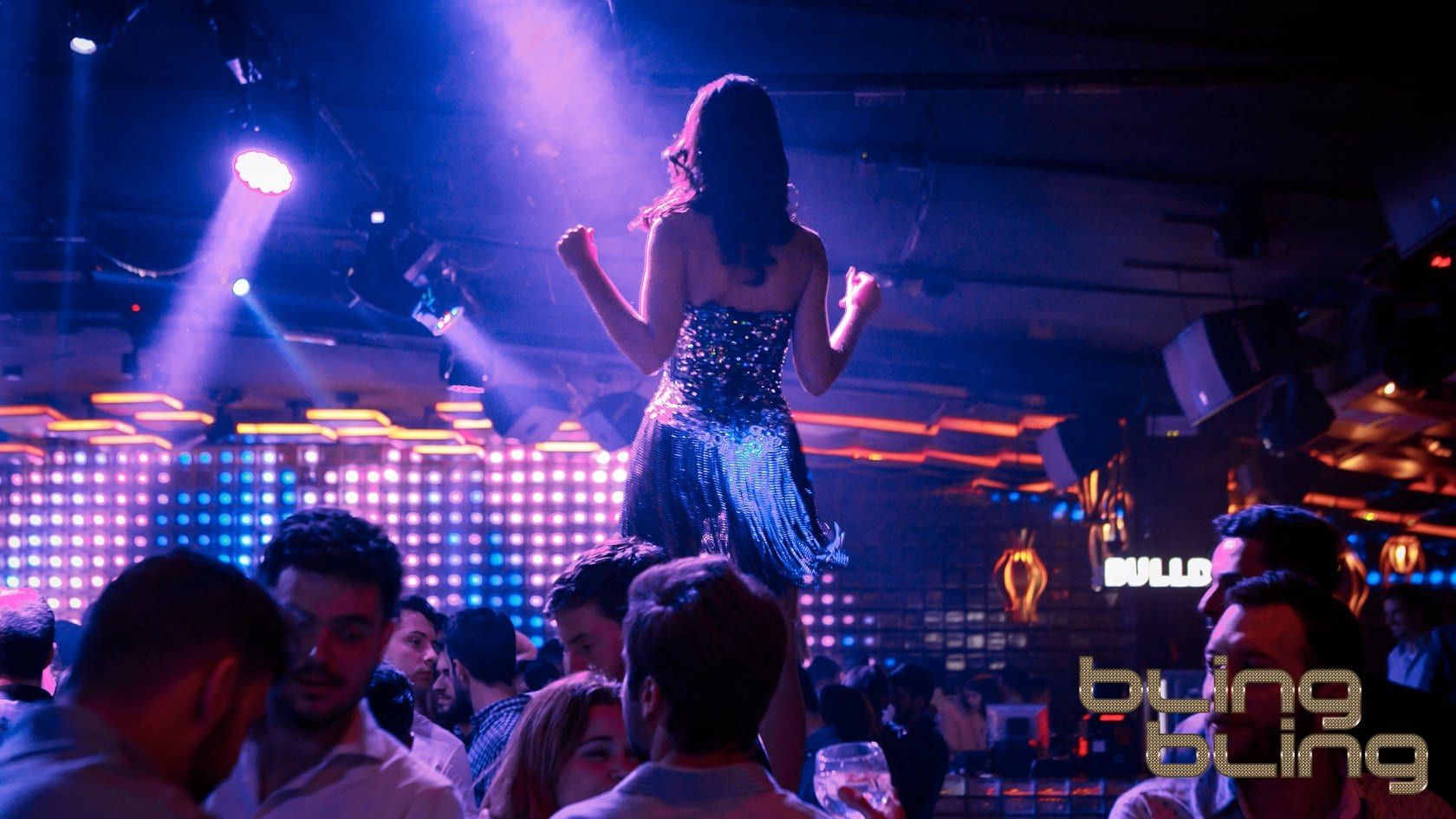 En la vibrante discoteca Bling Bling Barcelona, una mujer baila sobre una mesa de bar entre juerguistas entusiastas y luces radiantes. - ForTwo - Planes para parejas
