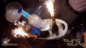 Bling Bling Barcelona presenta botellas adornadas con bengalas en un cubo de hielo brillante, rodeadas de una multitud enérgica inmersa en la vibrante atmósfera del club. - ForTwo - Planes para parejas