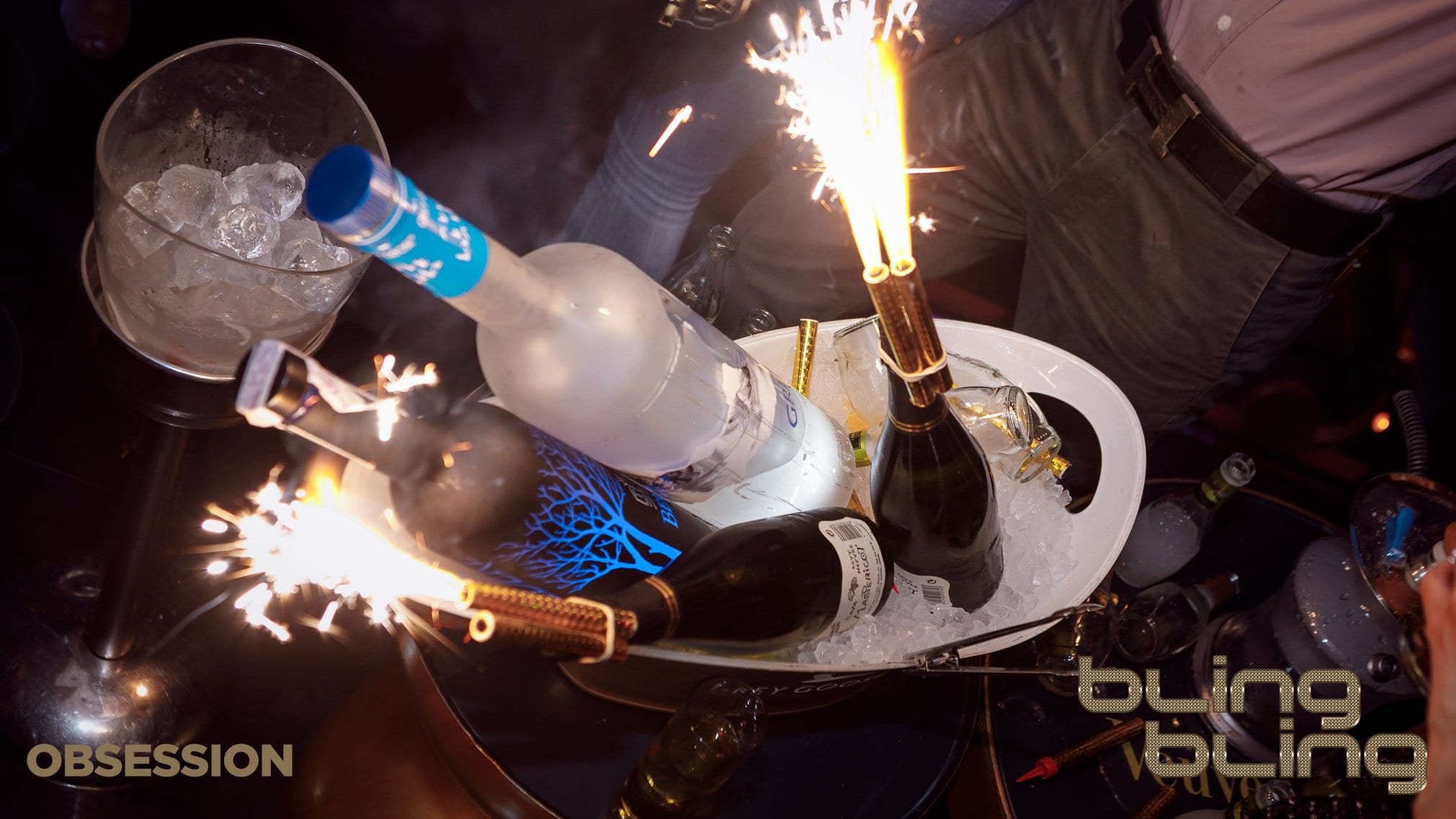 Bling Bling Barcelona presenta botellas adornadas con bengalas en un cubo de hielo brillante, rodeadas de una multitud enérgica inmersa en la vibrante atmósfera del club. - ForTwo - Planes para parejas
