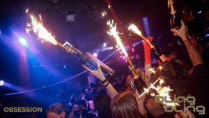 En el animado ambiente de Bling Bling Barcelona, los asistentes a la fiesta sostienen bengalas adheridas a botellas en medio de luces de colores. - ForTwo - Planes para parejas
