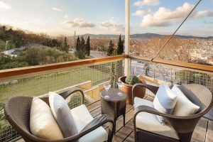 Disfrute de la serenidad del balcón del Hotel Miramar Barcelona GL, amueblado con dos sillas de mimbre y una mesa. Deléitese con las vistas del vibrante paisaje urbano de Barcelona y sus frondosos árboles bajo un cielo nublado, perfecto para una velada íntima. - ForTwo - Planes para parejas