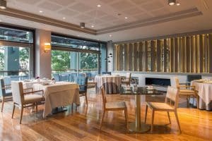 Elegante interior de restaurante con suelos de madera, mesas elegantes y grandes ventanales con vistas a los árboles exteriores, todo ello dentro del encantador ambiente del Hotel Miramar Barcelona GL (Barcelona). - ForTwo - Planes para parejas